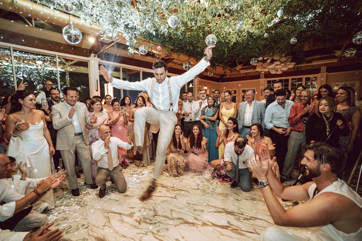 Groom dancing joyfully at wedding reception surrounded by cheering guests under disco ball and floral ceiling decor.