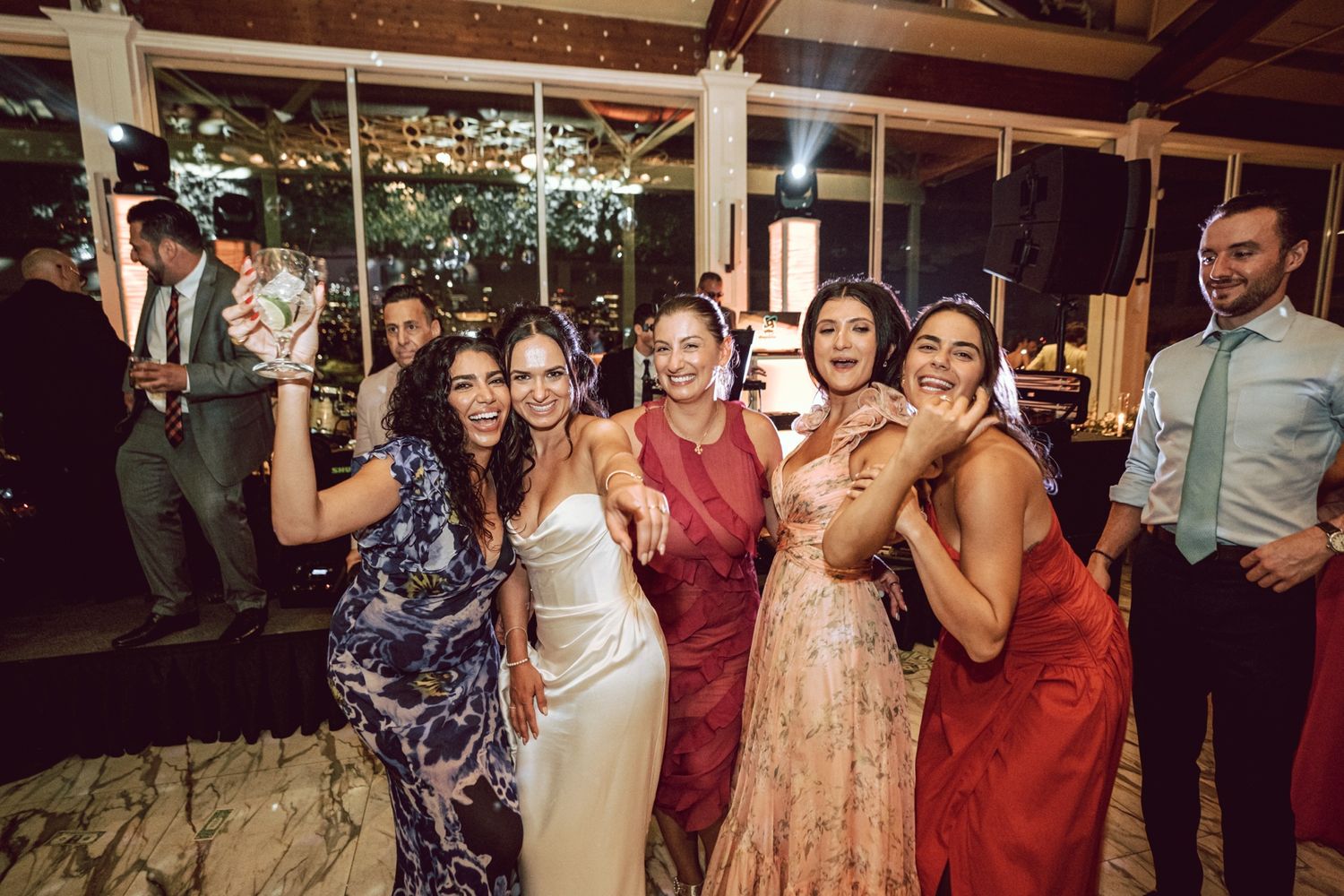 Five women in elegant dresses smile and pose together at a wedding reception.
