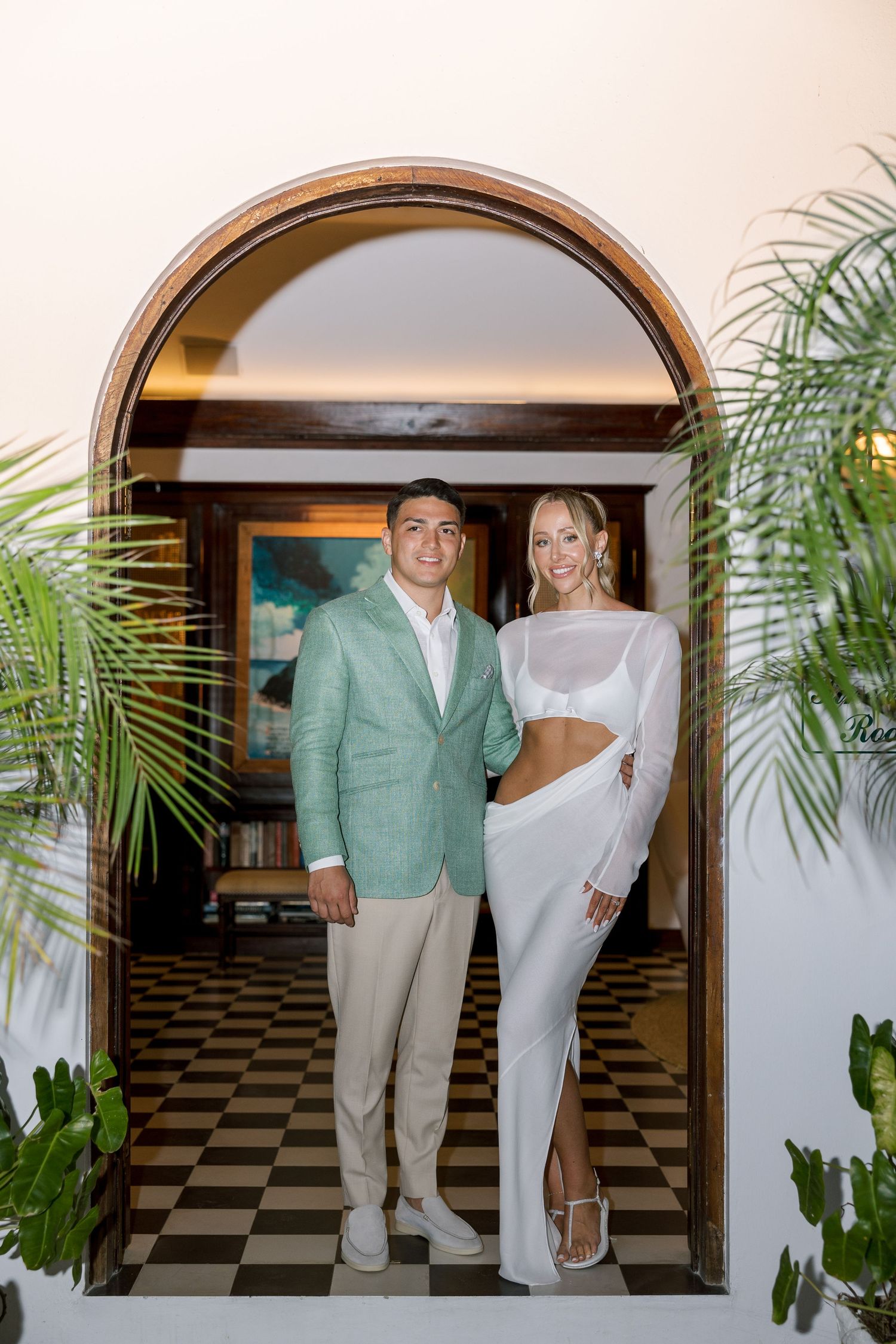 Couple posing in arched doorway, man in mint blazer and woman in white two-piece outfit, tropical venue.