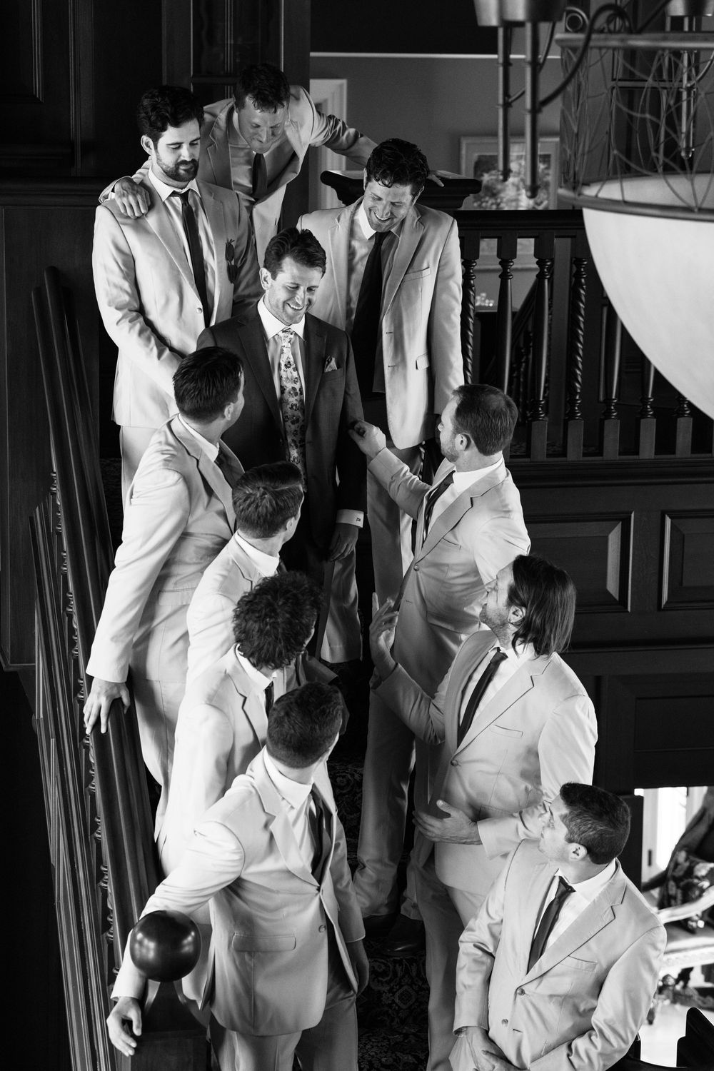 Groom surrounded by groomsmen in suits laughing together on staircase in black and white photo.