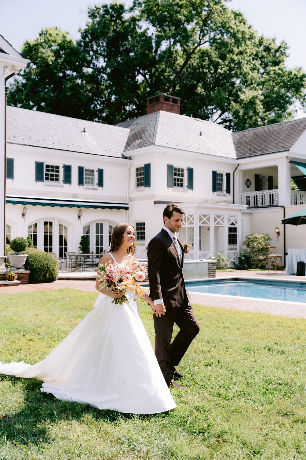 Bride and groom walk hand in hand on lawn of elegant white colonial estate with pool.