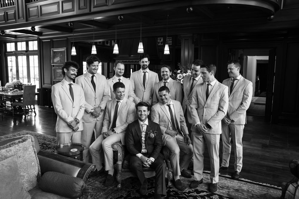 Black and white photo of a groom and groomsmen posing together in suits inside an elegant wood-paneled venue.