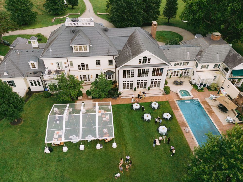 Aerial view of a luxury estate with a glass tent, swimming pool, and manicured lawn during an outdoor event.