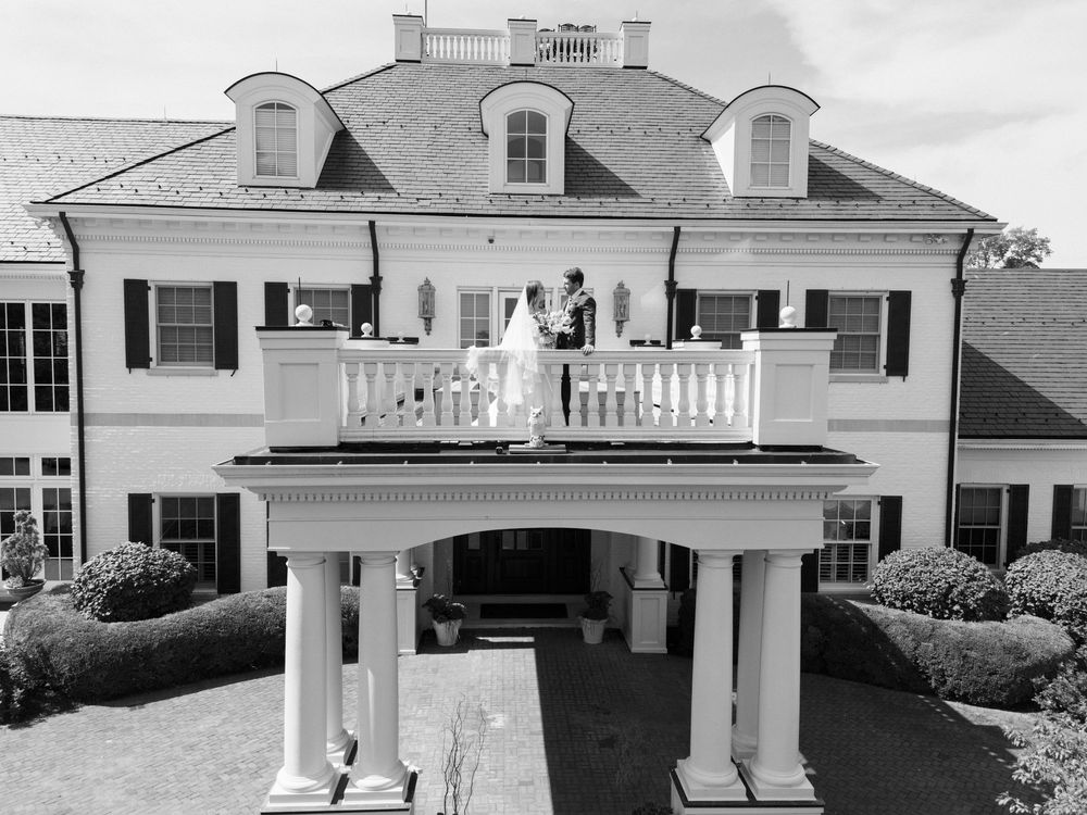 Black and white photo of a couple sharing a romantic moment on a grand estate balcony with white columns.