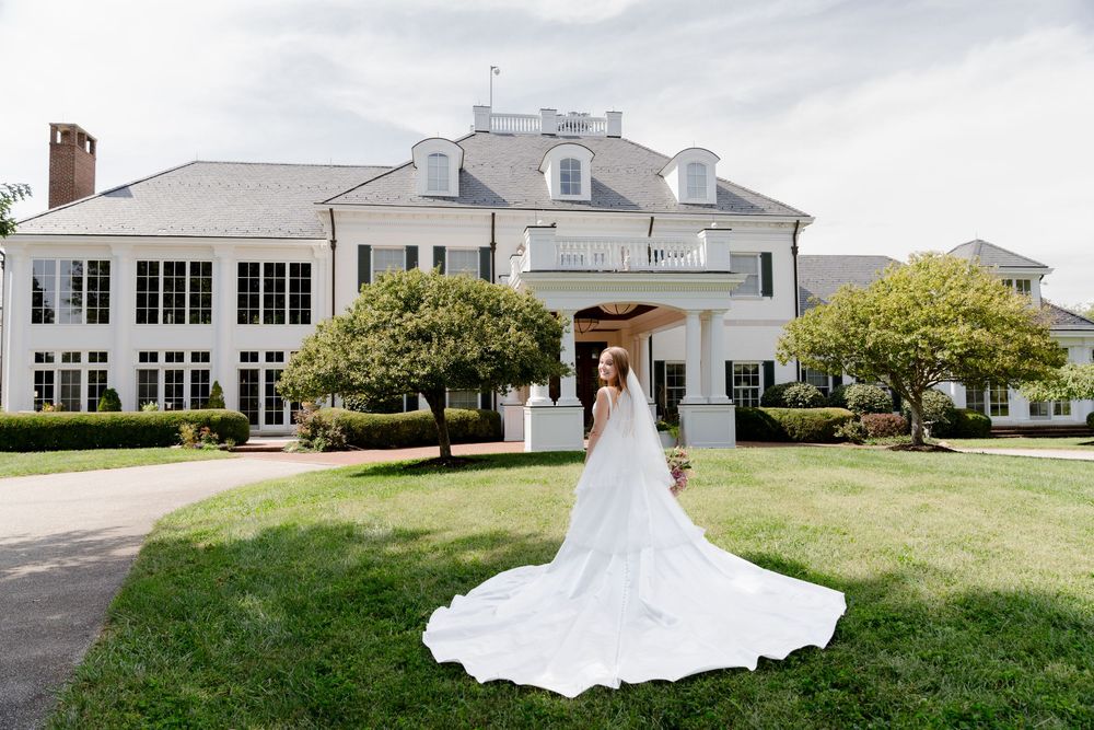 Bride in flowing white gown with long train stands on green lawn before elegant white colonial mansion.