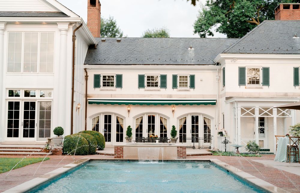 Elegant white mansion with green shutters, arched windows, and a serene outdoor swimming pool in a lush garden setting.