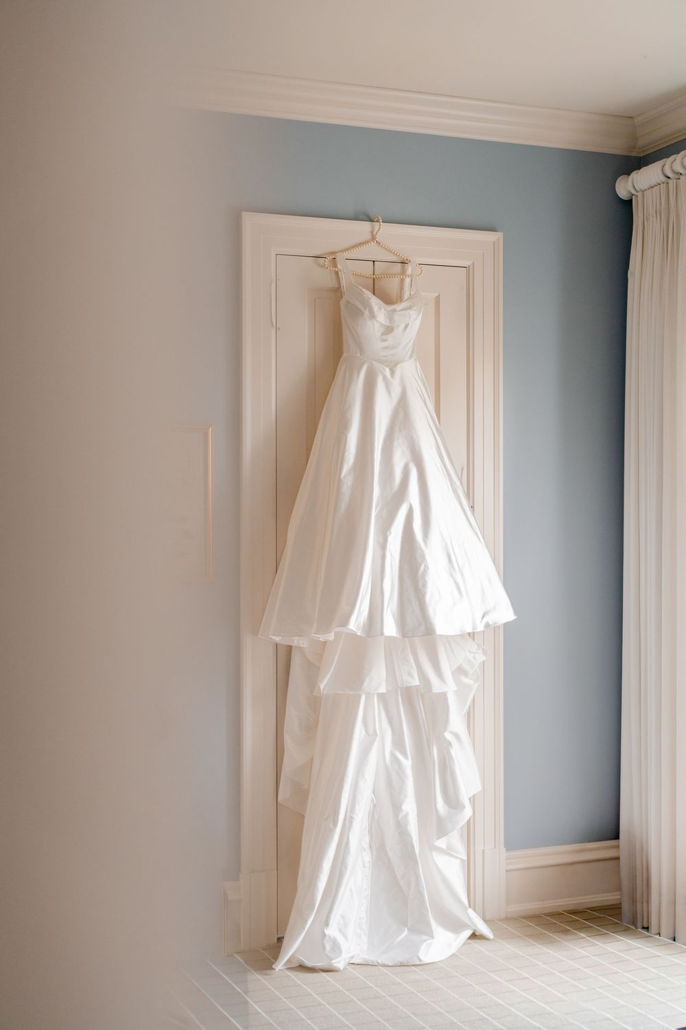 Elegant white wedding gown with tiered skirt hanging on a door in a soft blue and cream bridal suite.