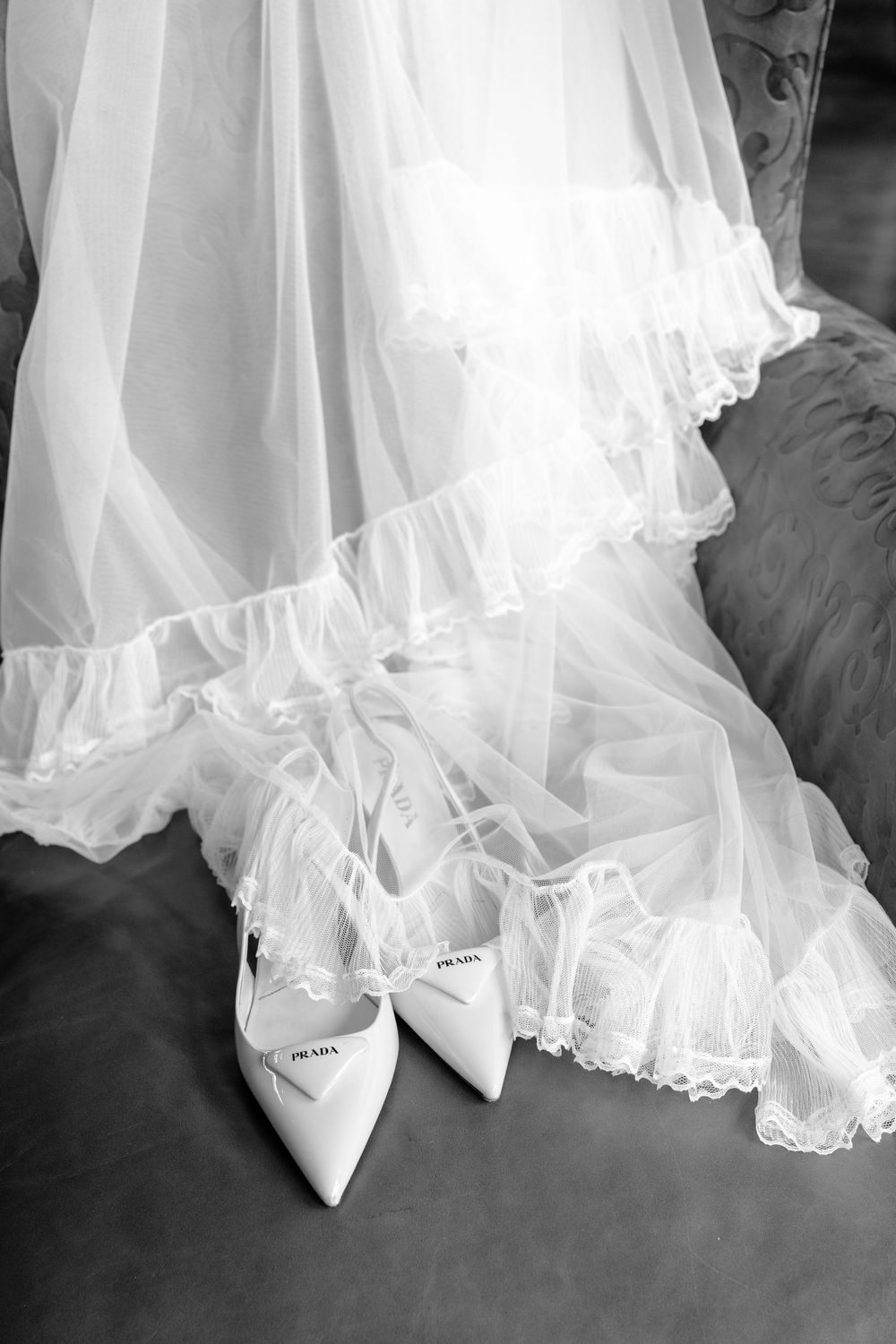 Black and white photo of Prada bridal heels resting beside a flowing lace wedding dress on a velvet chair.