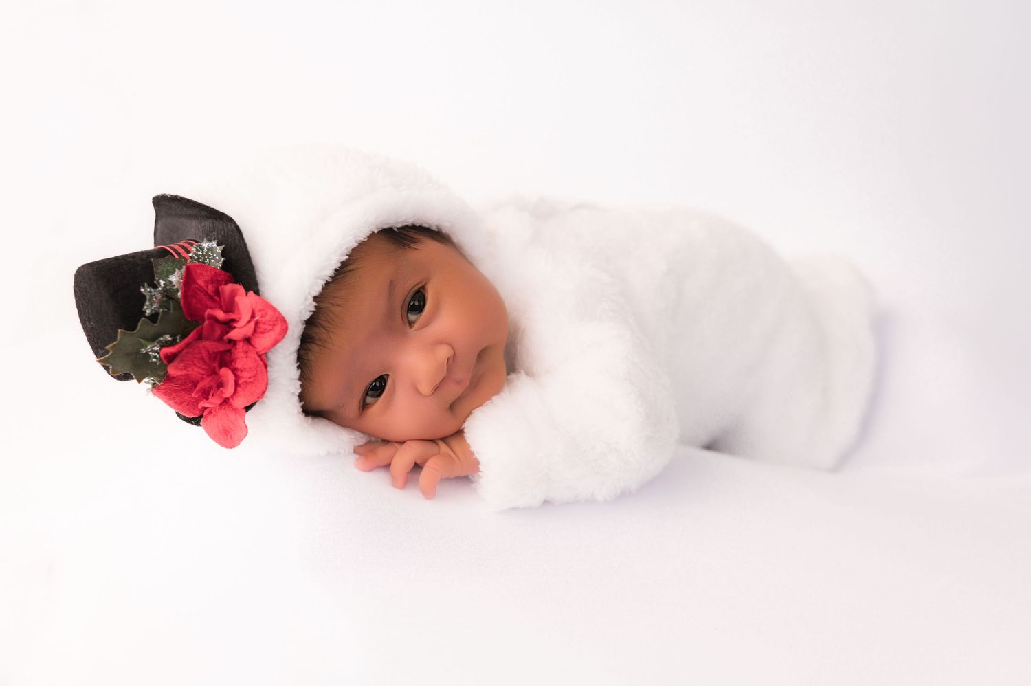 Newborn baby in white fluffy outfit with a black hat adorned with red flowers, posed on white background.