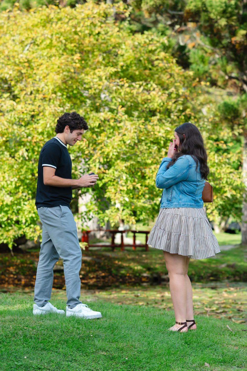 Young couple in a park, man showing phone to surprised woman in denim jacket and plaid skirt outdoors.