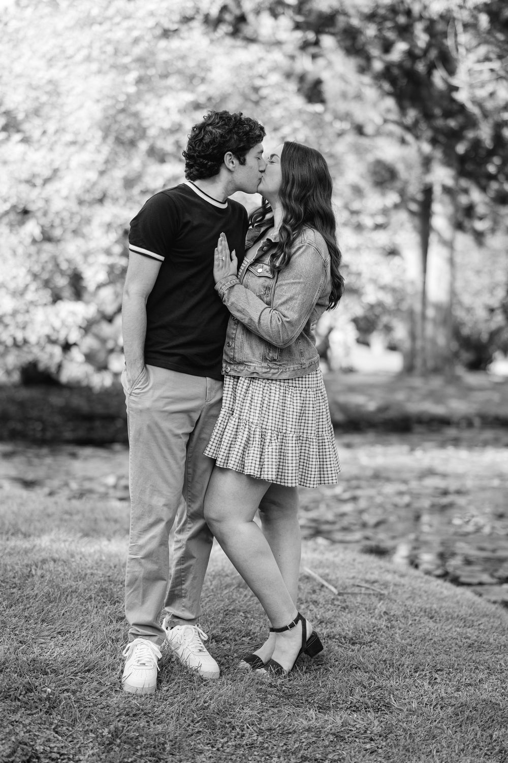 Black and white photo of a couple kissing in a park, woman in plaid skirt and denim jacket, man in black polo shirt.