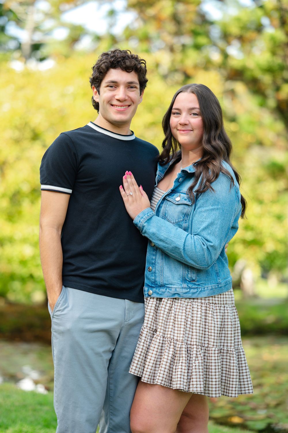 A smiling young couple poses together outdoors in a lush green park, wearing casual summer outfits.