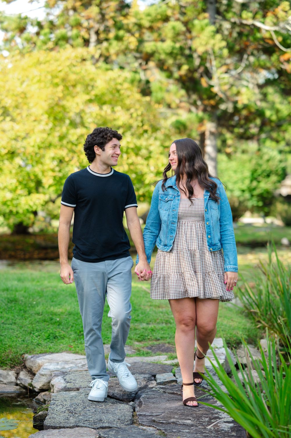 Young couple holding hands and smiling at each other while walking on stone path in lush green park.