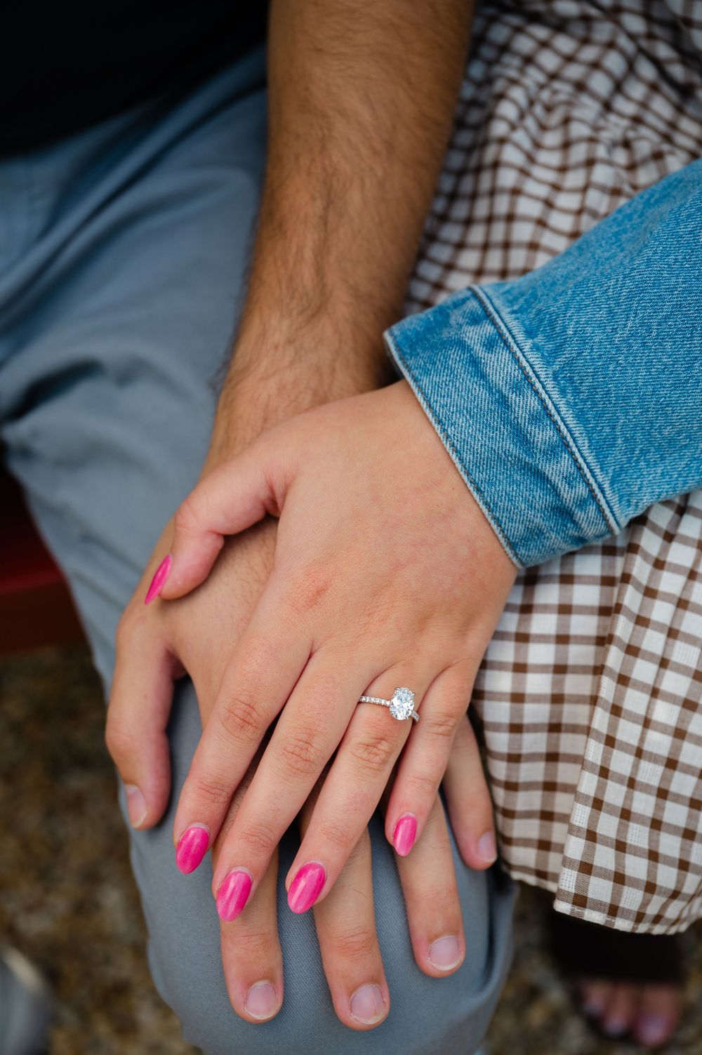 Close-up of an engagement ring with oval diamond on womans hand with pink nails resting on mans knee.