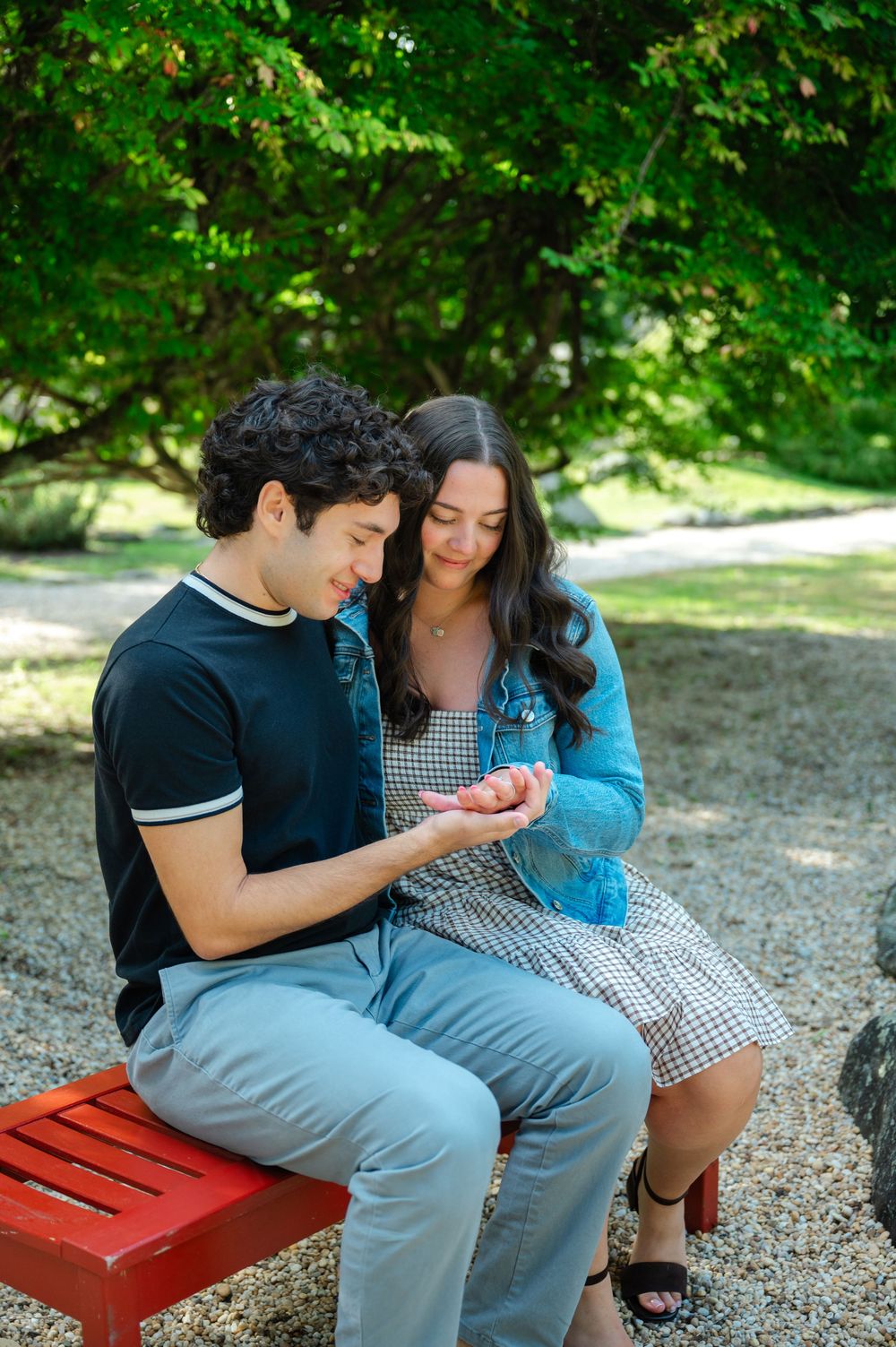 Couple sitting on a red bench in a park, looking at something in their hands, surrounded by lush green trees.