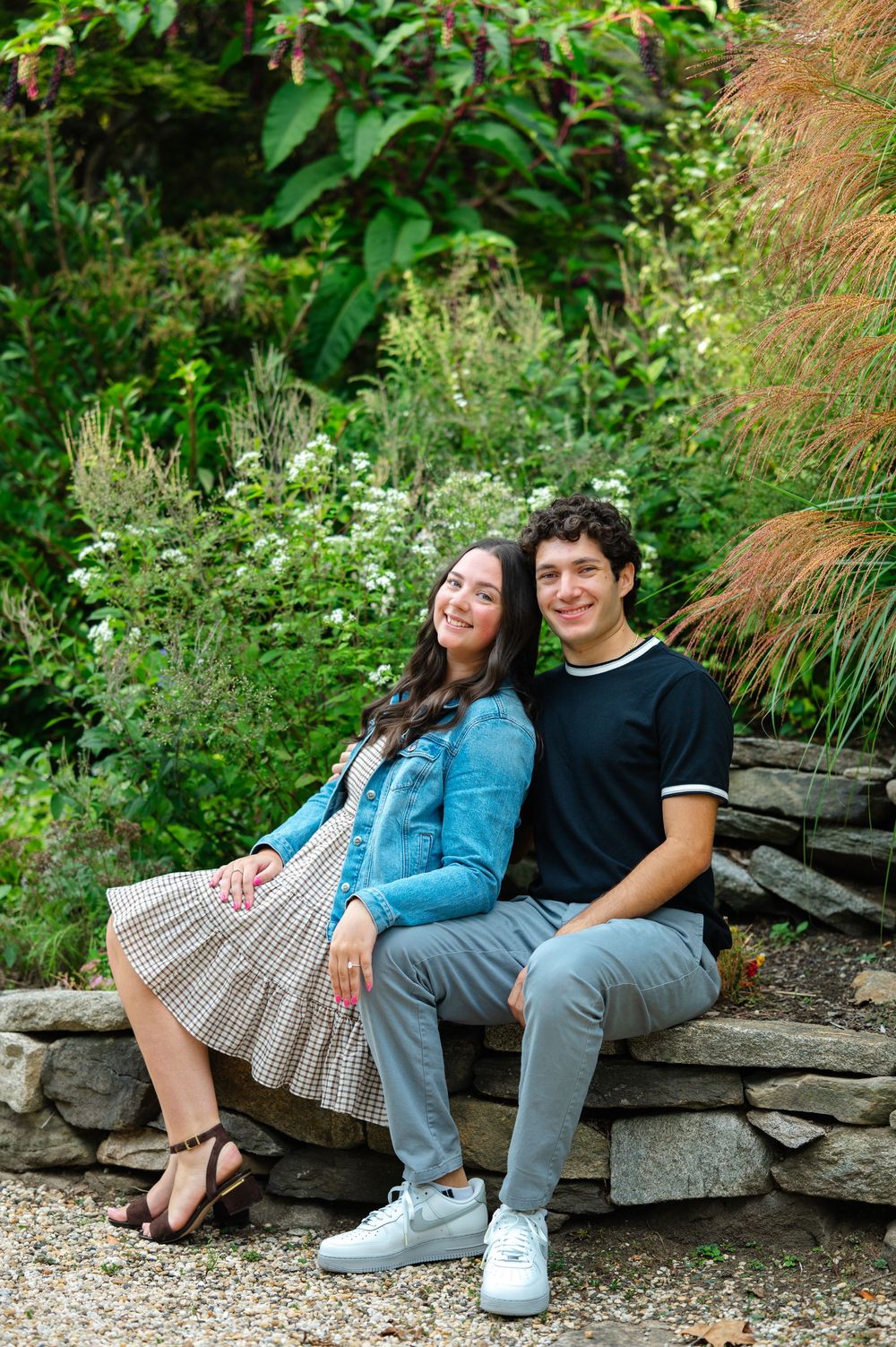 A young couple smiles while sitting together on a stone bench surrounded by lush green garden foliage.
