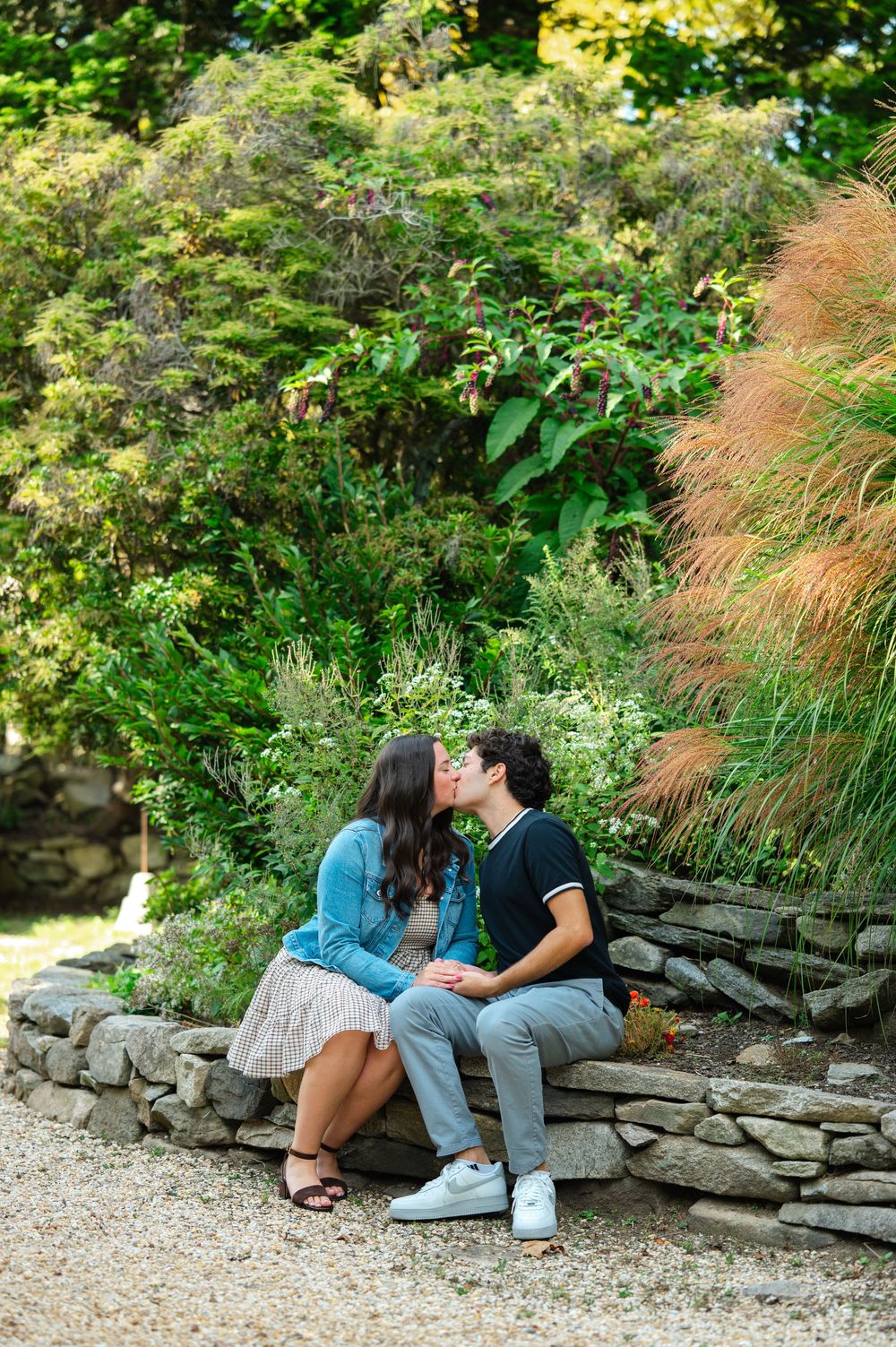A couple shares a romantic kiss while sitting on a stone wall surrounded by lush green garden foliage.