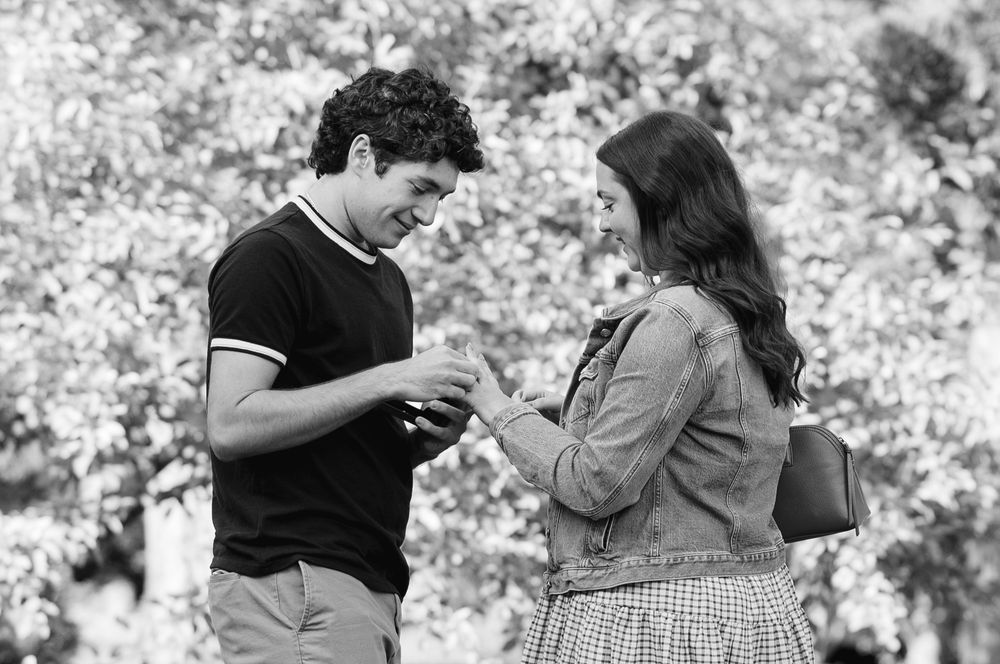 Black and white photo of a young couple sharing a moment outdoors, with blooming trees in the background.