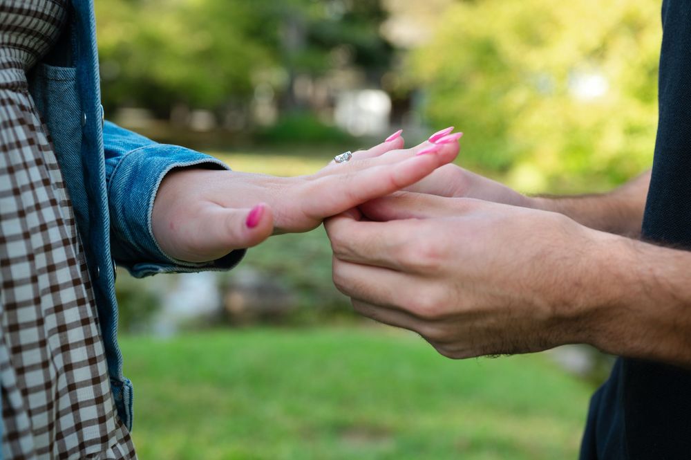 Close-up of a man placing a ring on a woman's finger outdoors in a green park setting.