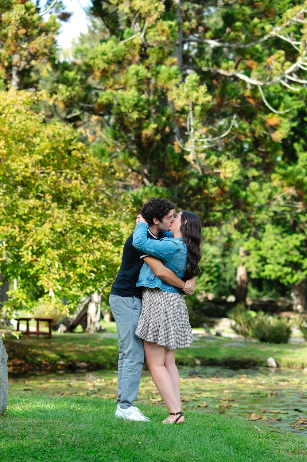 Couple sharing a romantic kiss in a lush green park surrounded by trees on a sunny day.