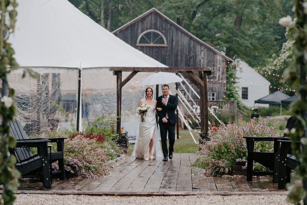 Bride and groom walking hand-in-hand on a garden path with a rustic barn and white tent backdrop.