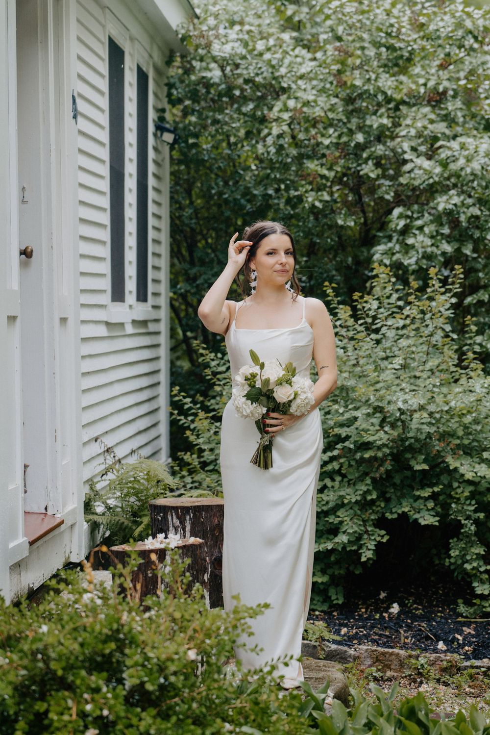 Bride in white satin gown holding floral bouquet stands beside a white building surrounded by lush greenery.