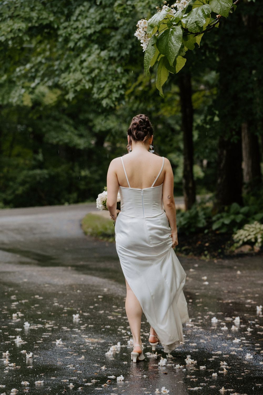 Bride in white satin slip dress walks rain-wet path holding bouquet, back view, lush green trees.