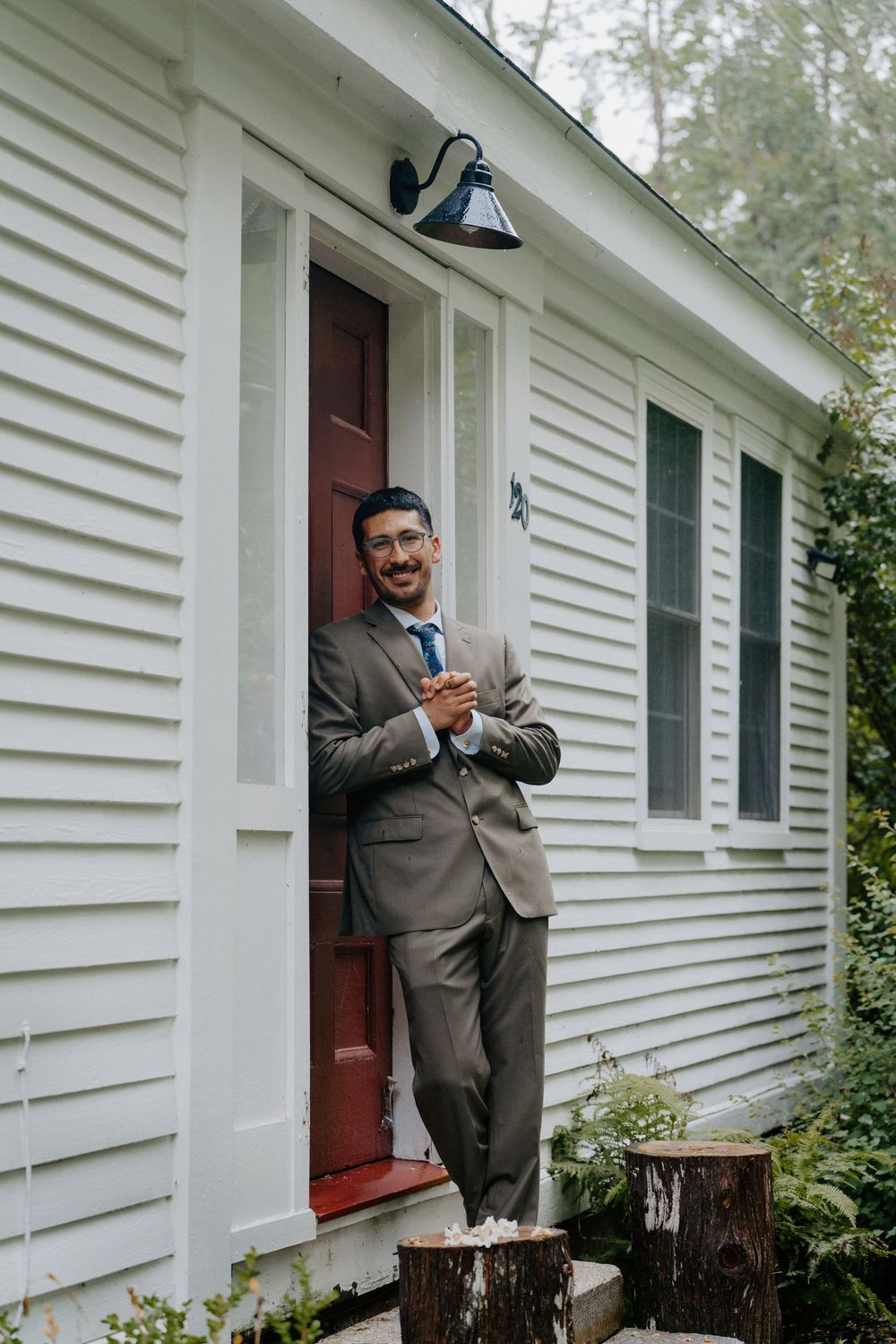 Smiling groom in gray suit and blue tie leaning against red door of white clapboard house.