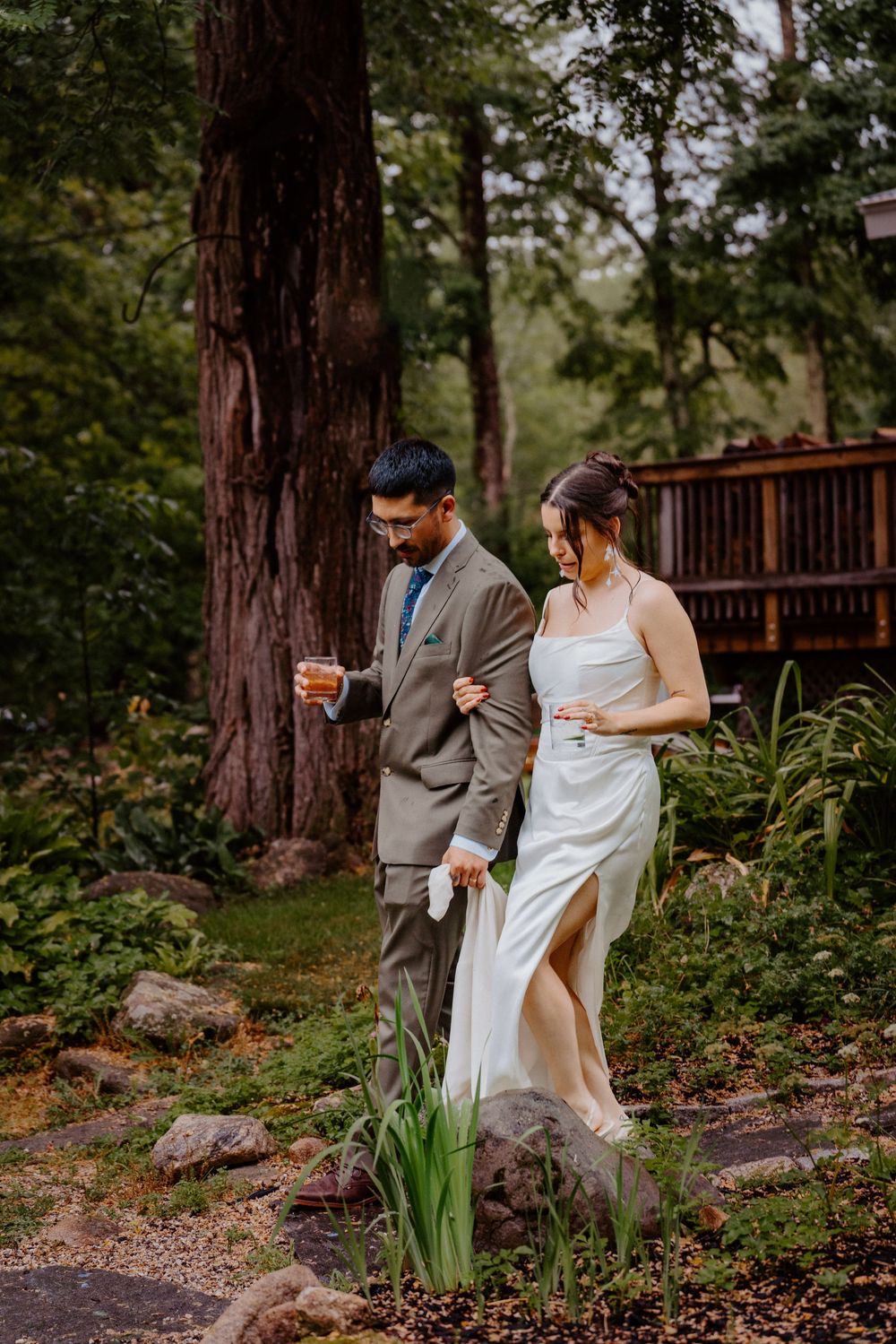 Bride and groom walking through lush garden setting, groom in tan suit holding drink, bride in elegant white gown.