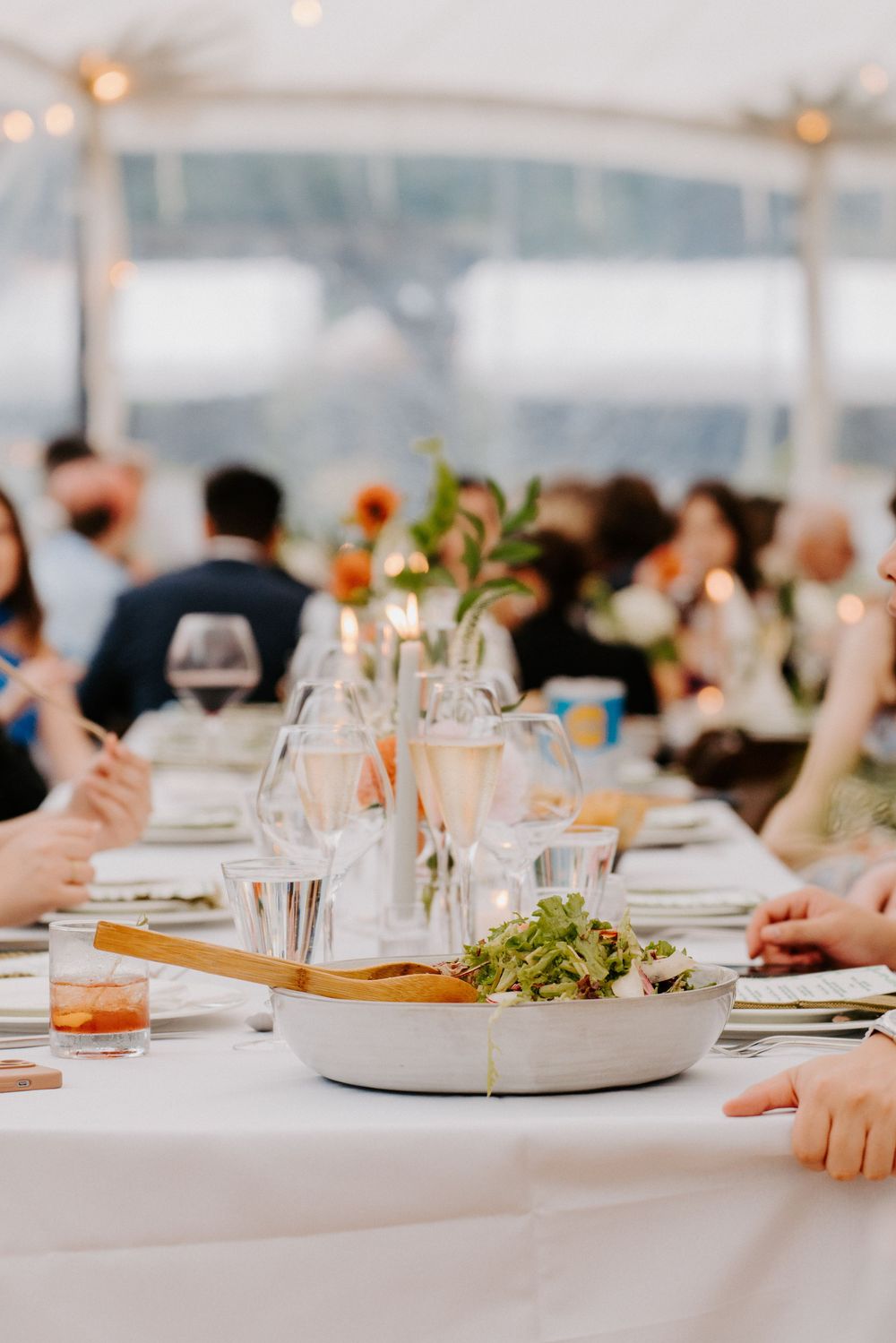 Elegant wedding reception table with salad bowl, wine glasses, candles, and floral centerpieces under a white tent.