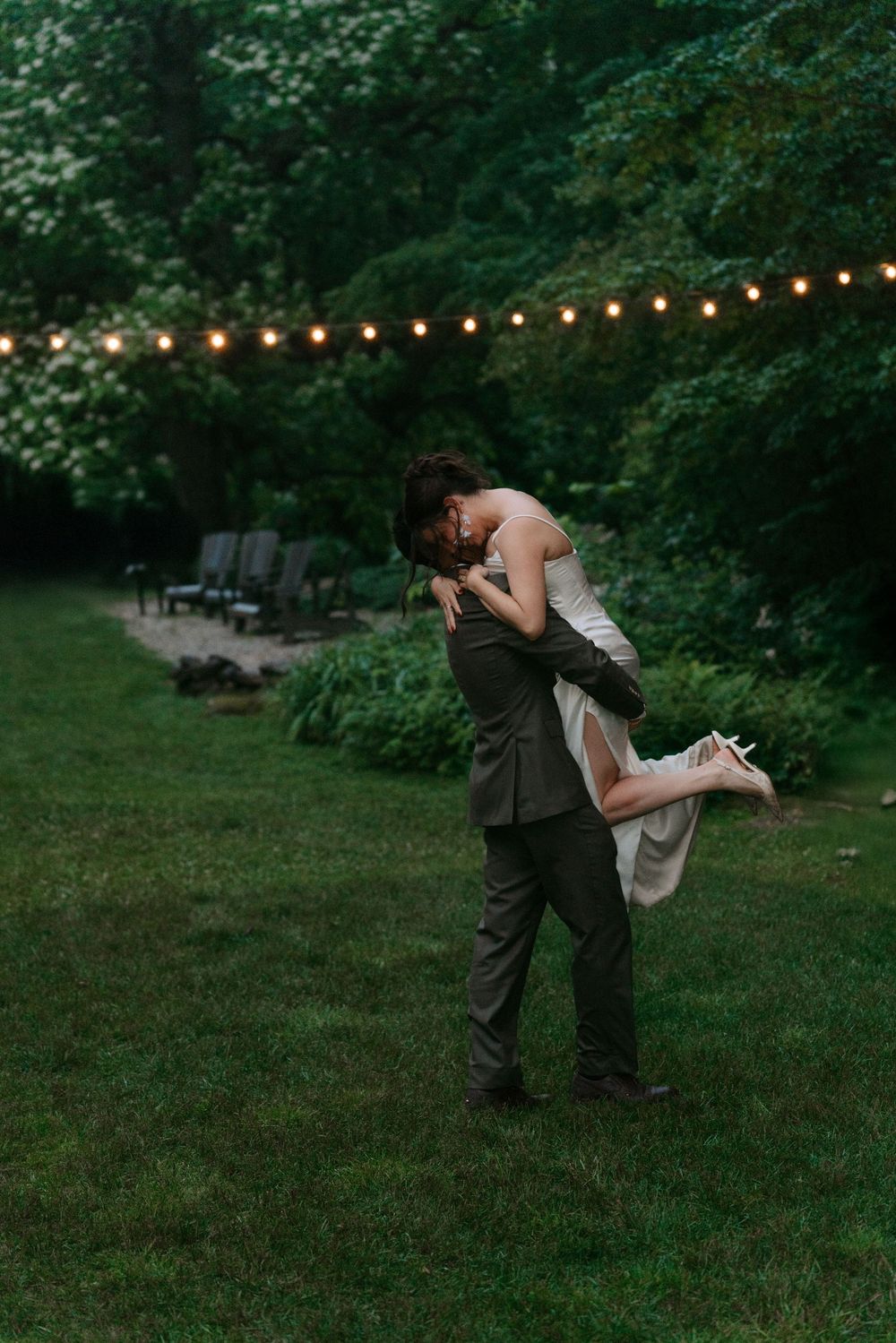 Groom lifts bride in a romantic embrace on lush green lawn with string lights and trees at dusk.