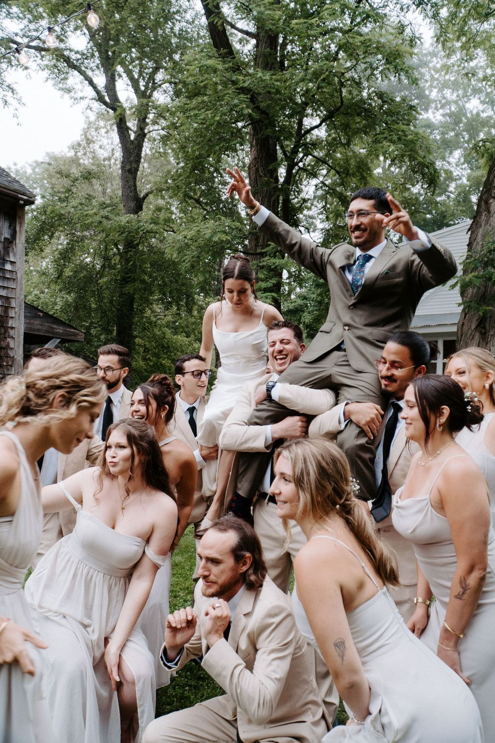 Joyful wedding party in white and tan attire lifting the bride and groom outdoors among lush green trees.