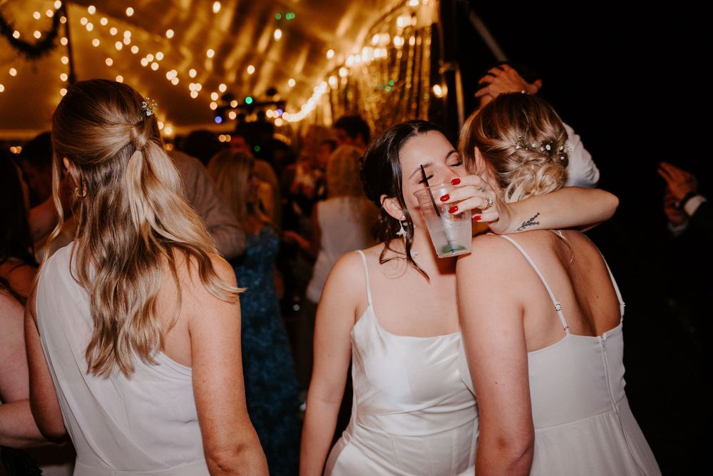 Women in white dresses dancing and embracing at a wedding reception under warm string lights.