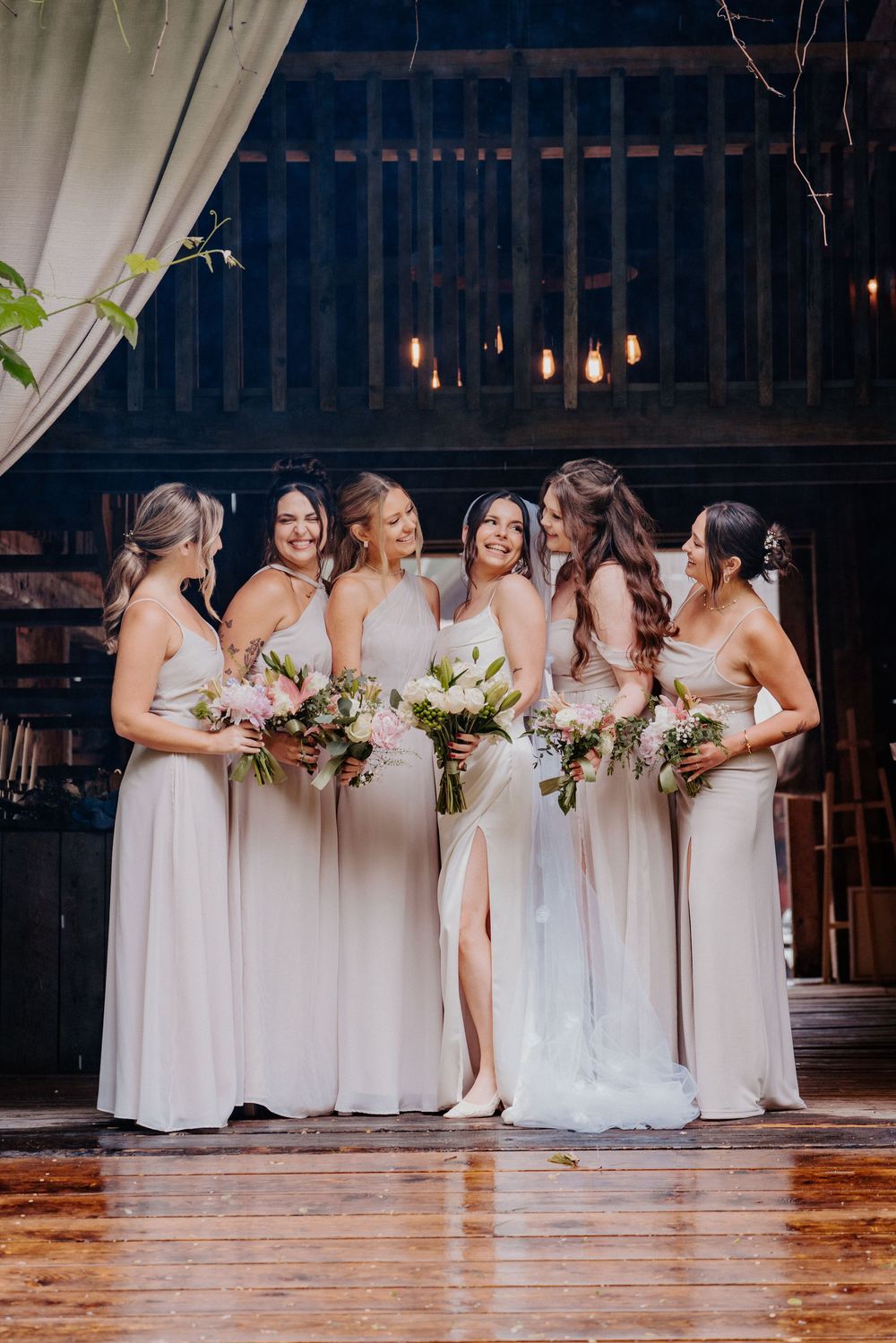Bride and five bridesmaids in champagne gowns holding bouquets inside a rustic barn venue.
