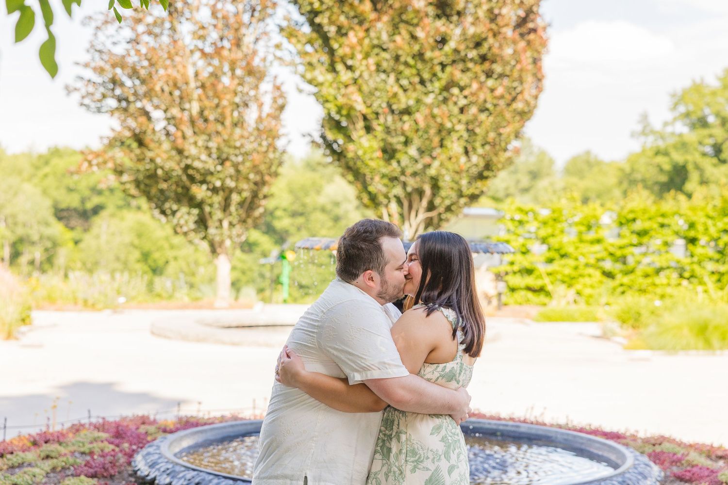 Couple sharing a romantic kiss near a decorative fountain surrounded by lush green trees and colorful flowers outdoors.