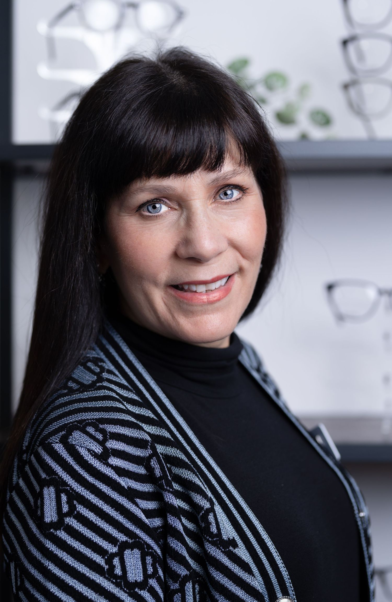 Professional woman with dark hair and blue eyes smiling in front of eyeglass display shelves.