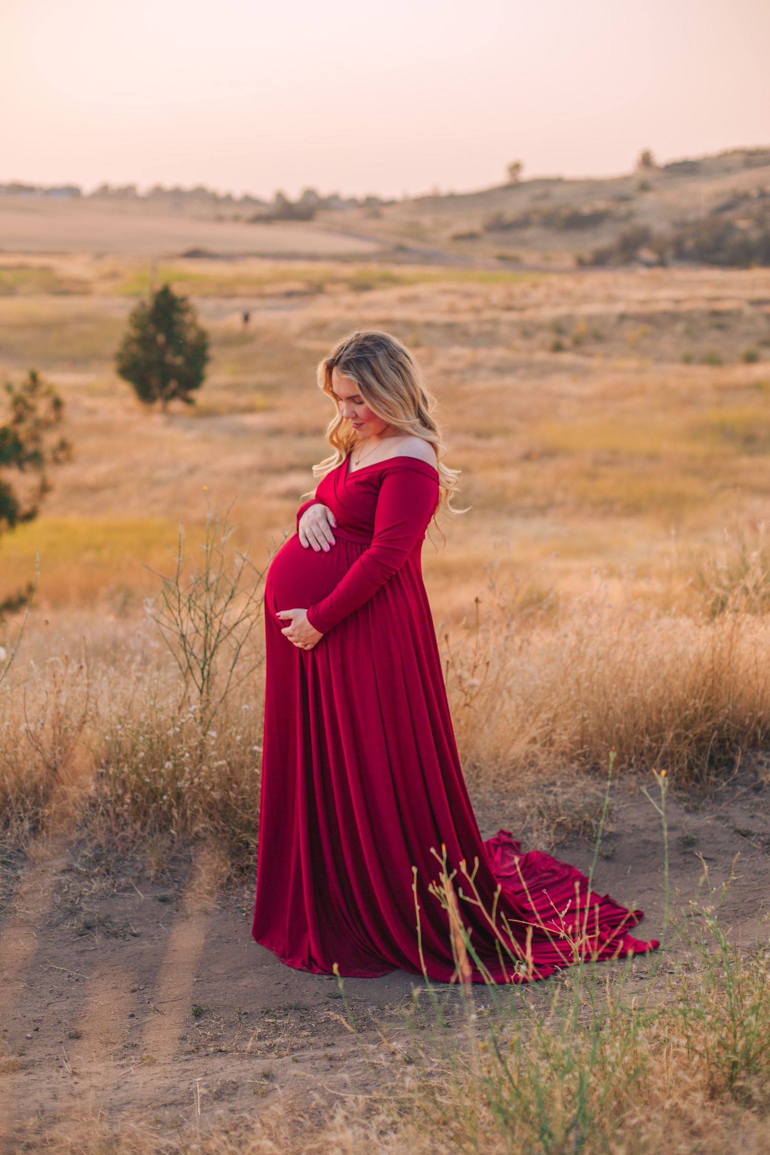 Pregnant woman in elegant red off-shoulder gown cradling baby bump in golden field at sunset.