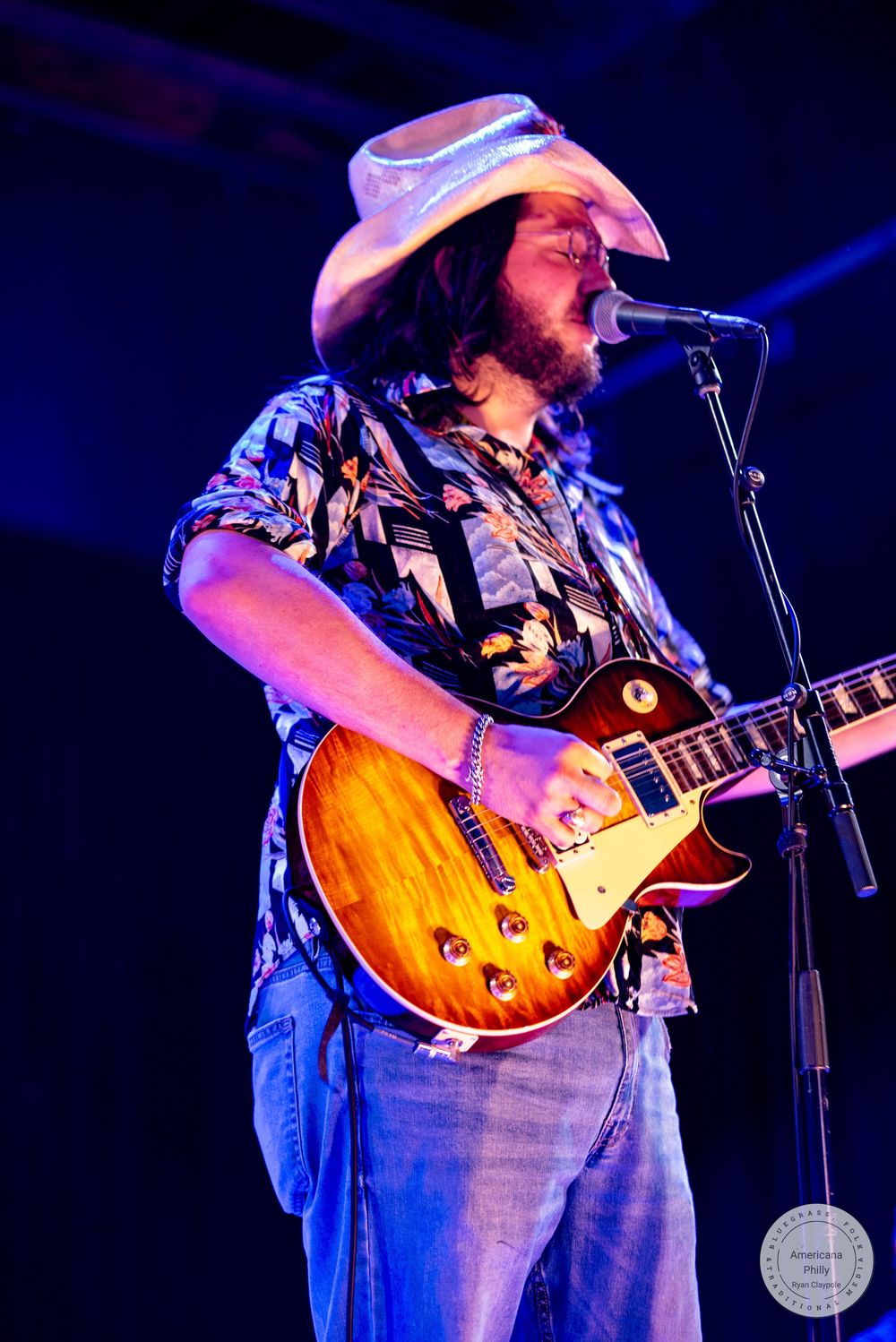 Bearded musician in white cowboy hat plays sunburst Les Paul guitar while singing into microphone on stage.