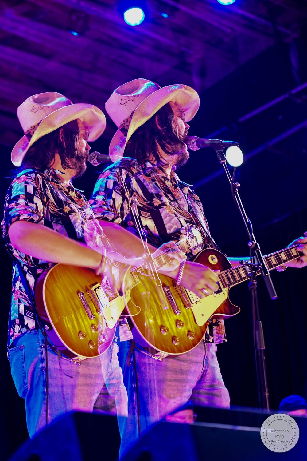 Two identical twin guitarists in cowboy hats perform on stage with Gibson Les Paul guitars under purple lights.