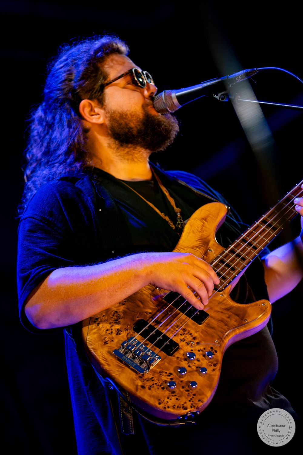 Bearded male musician with long hair playing a wood grain bass guitar on stage under blue and purple lights.
