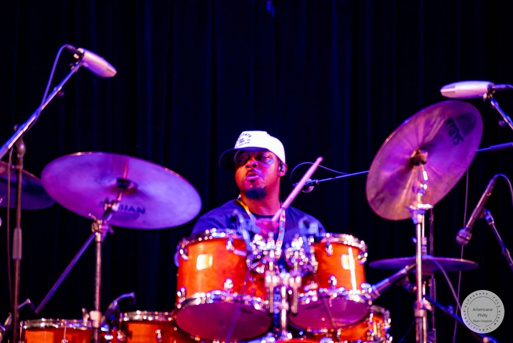 A drummer performs on stage with orange drums and cymbals under purple stage lighting.