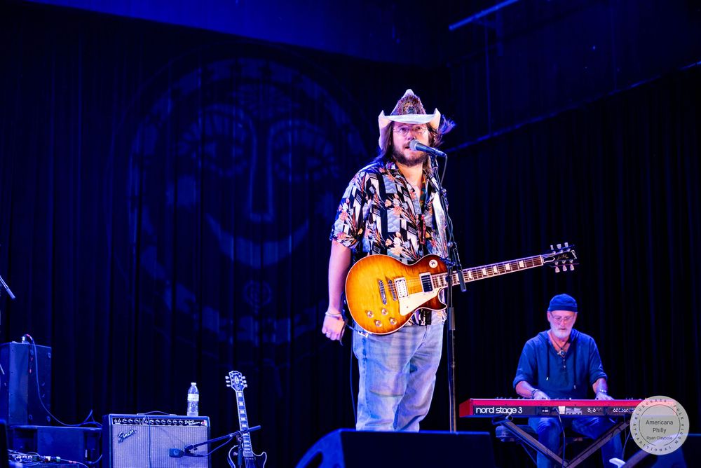 Country musician in cowboy hat plays Les Paul guitar on stage with keyboardist performing live at a music venue.