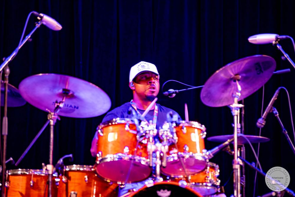 A drummer performing on stage with orange drums and cymbals under blue stage lighting.