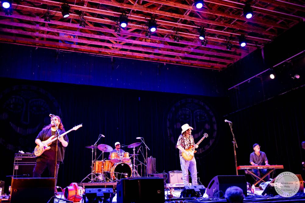 Live band performing on stage with guitars, drums, and keyboards under colorful stage lights at an outdoor venue.