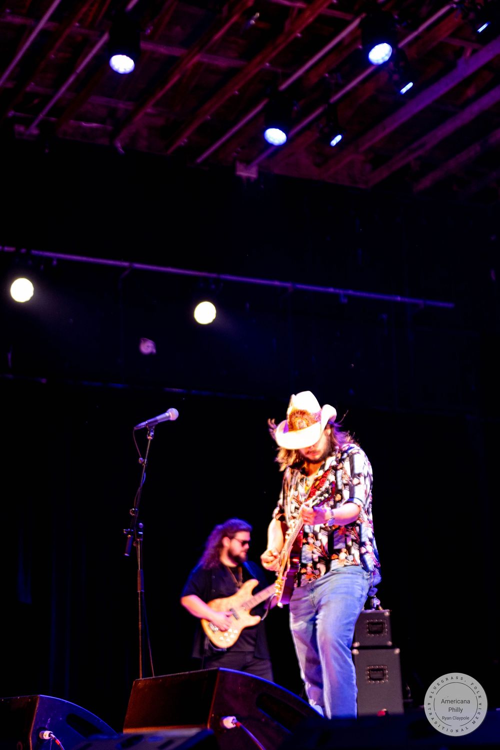 Two musicians perform on stage, one in a cowboy hat playing guitar under colorful concert lights.