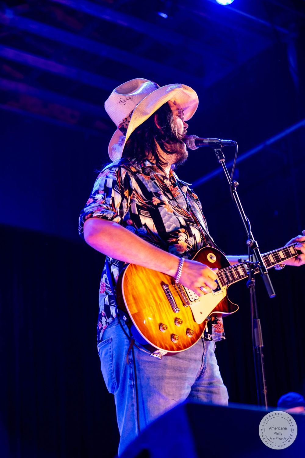 Guitarist in cowboy hat and floral shirt plays Les Paul guitar on stage under blue lights.
