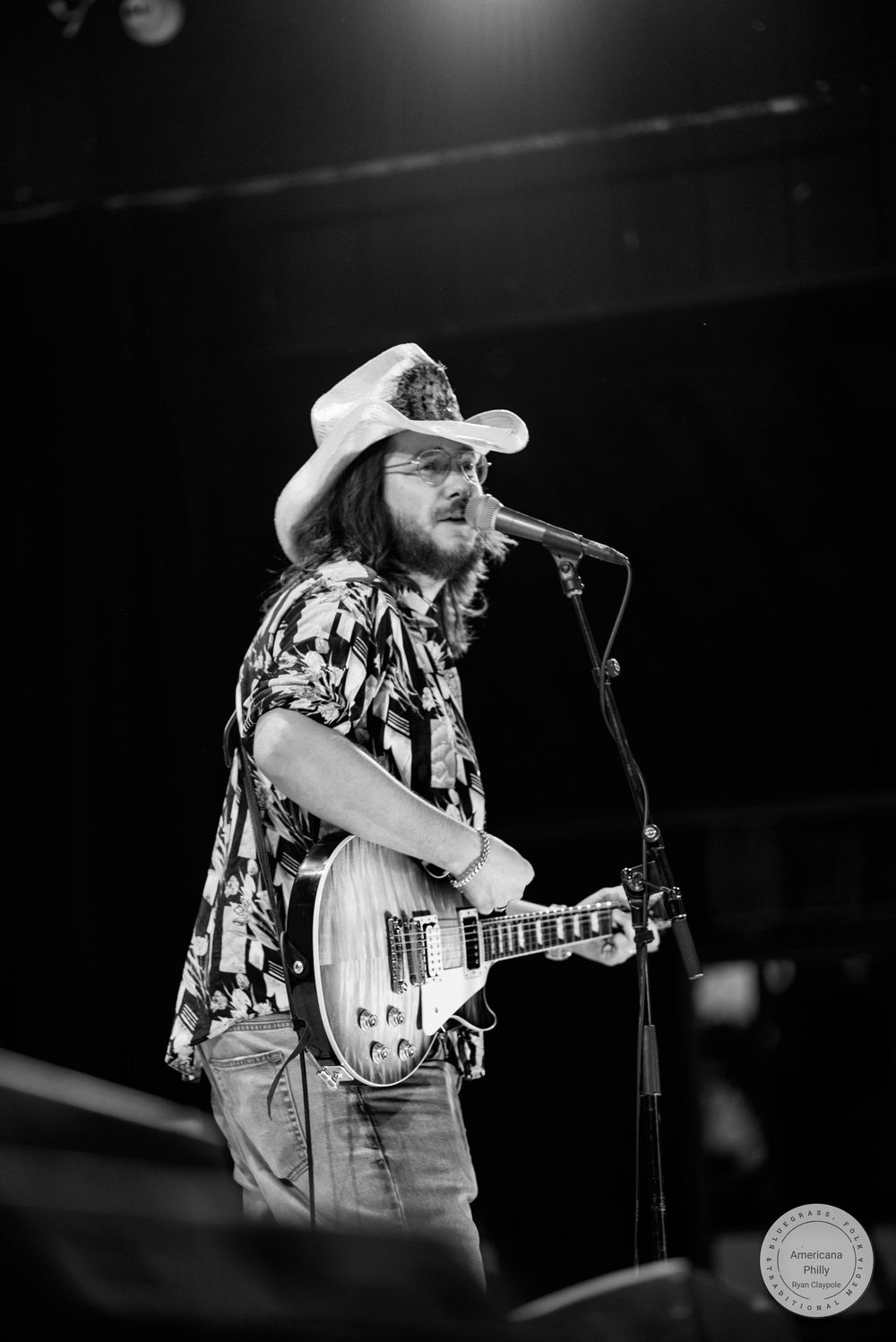 Black and white photo of a male musician in cowboy hat playing electric guitar and singing into microphone on stage.