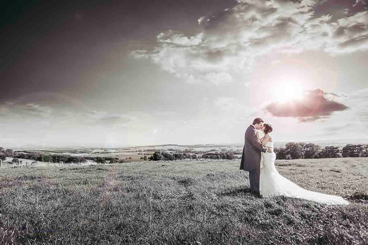 A bride and groom embrace in a sweeping countryside field under dramatic cloudy skies at Roundthorn Country House -Wedding venue -Penrith