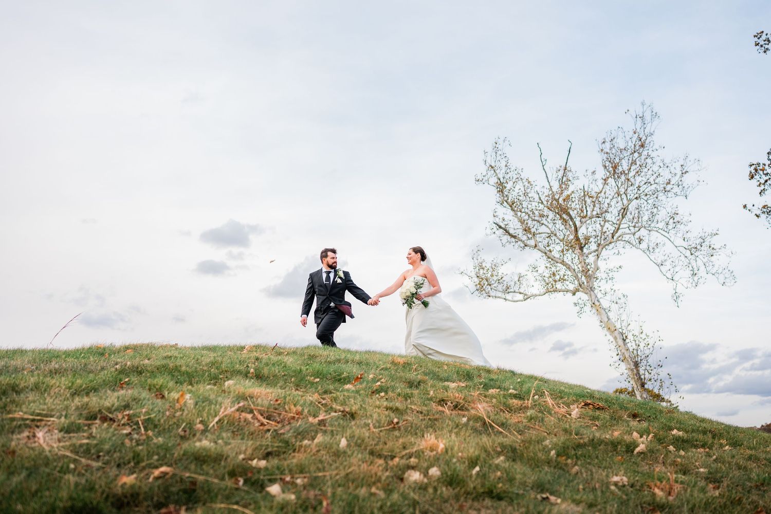 Bride and groom holding hands running on a grassy hill with wedding dress flowing in the wind on a cloudy day at Great River Golf Club