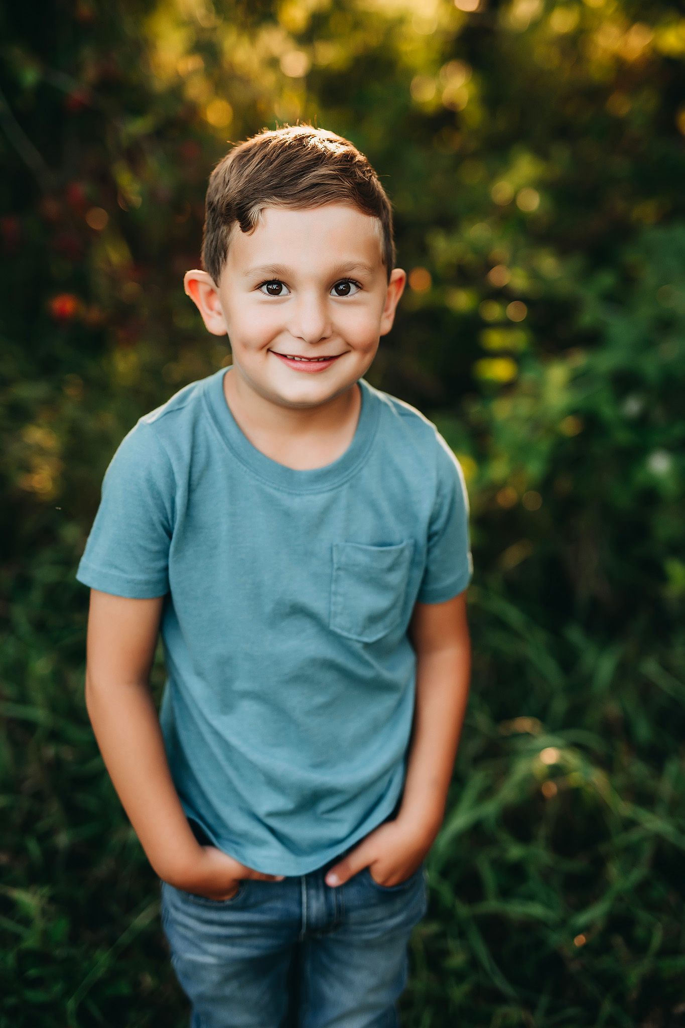 Smiling young boy in blue t-shirt and jeans standing outdoors with green foliage background.